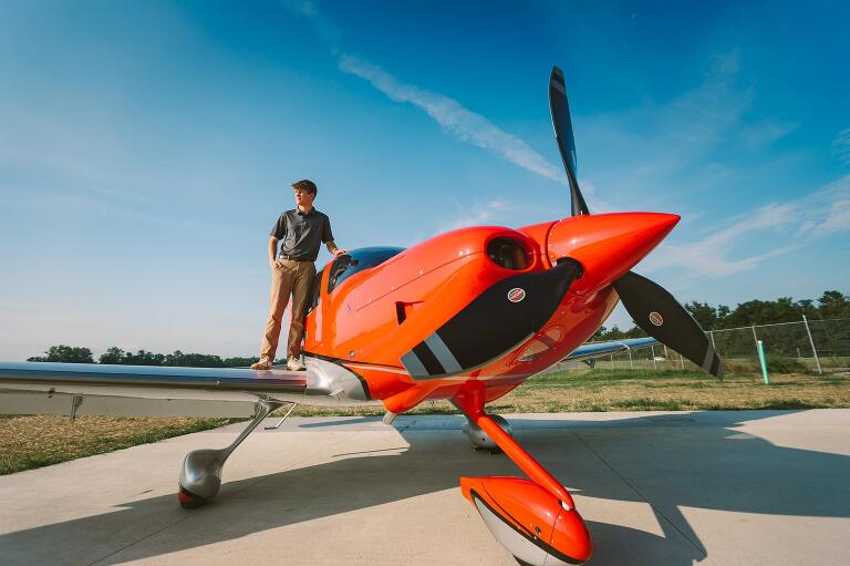 Senior guy standing on the wing of a vibrant red airplane against a bright blue sky at Dayton Wright Brothers Airport.