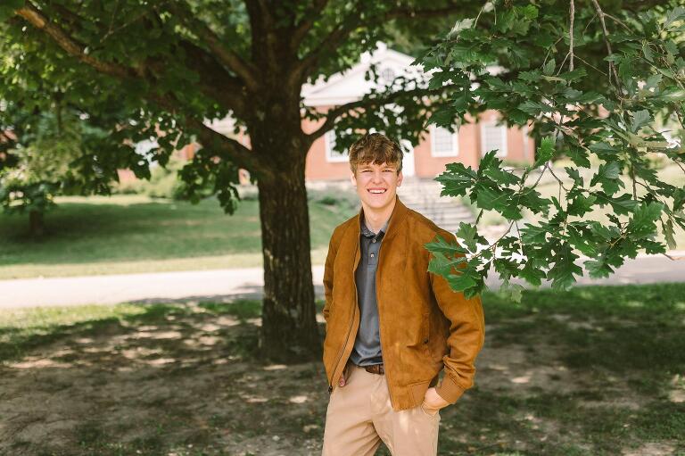 Senior guy wearing a brown leather bomber jacket posing under a large oak tree at Carillon Park.